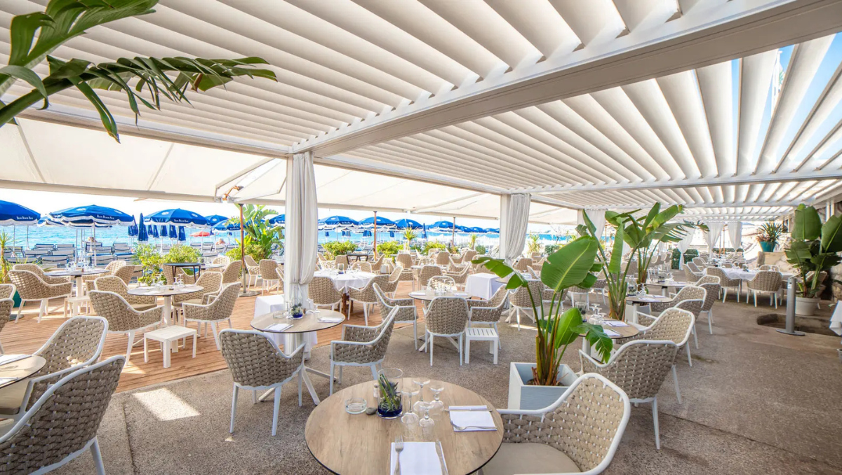 Luxury Coastal Pergola Dining Experience Open-air beachfront restaurant seating area featuring a modern white pergola with adjustable louvers, woven dining chairs, tropical plants, and ocean views in the background.